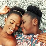 From above of positive African American female friends with closed eyes lying on stony beach while touching foreheads in summer