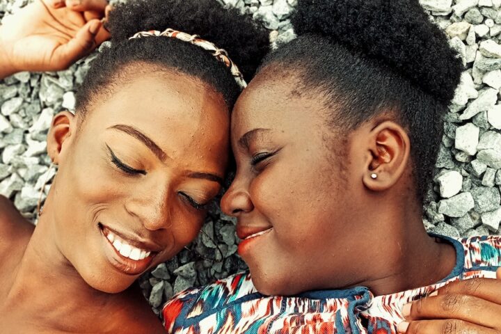 From above of positive African American female friends with closed eyes lying on stony beach while touching foreheads in summer