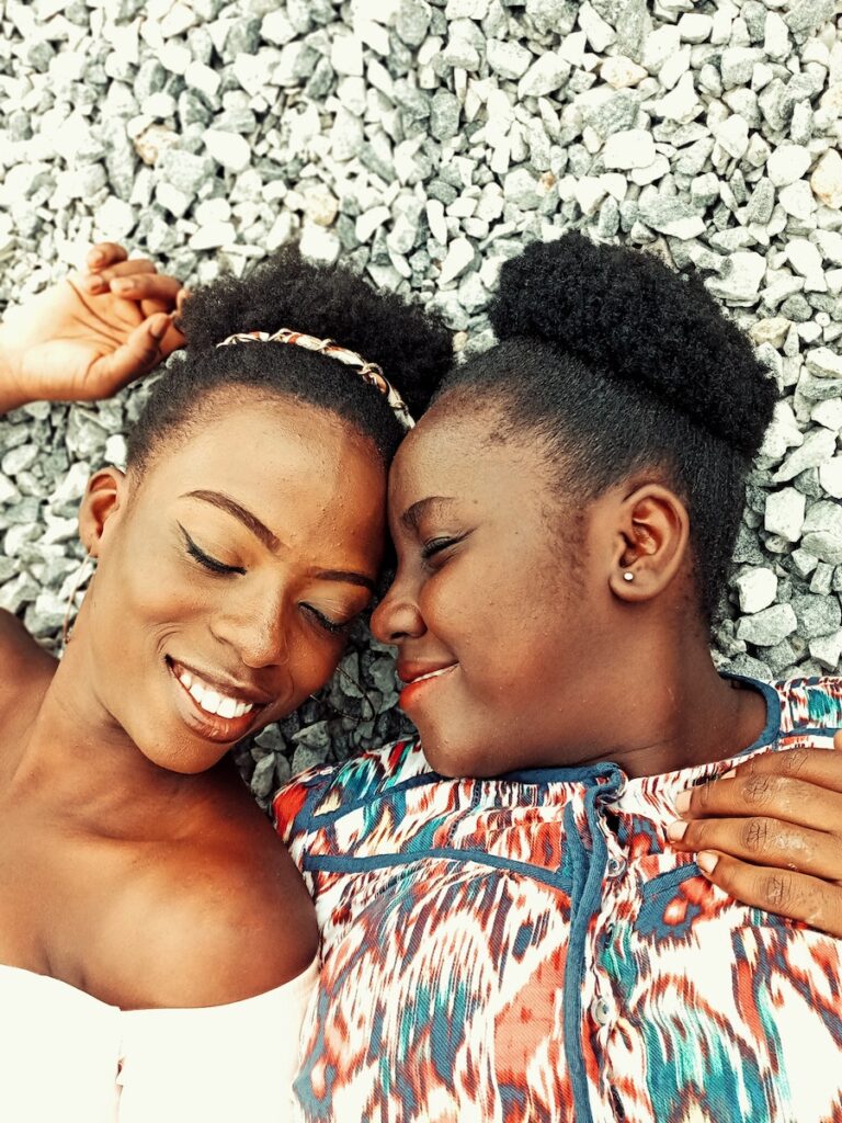 From above of positive African American female friends with closed eyes lying on stony beach while touching foreheads in summer