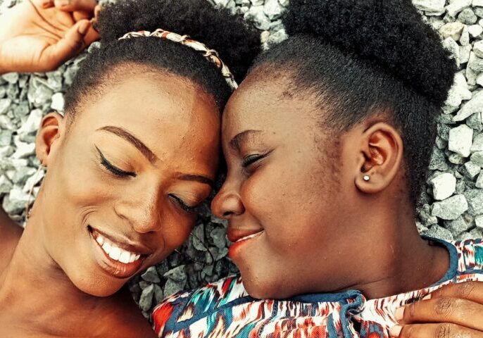From above of positive African American female friends with closed eyes lying on stony beach while touching foreheads in summer